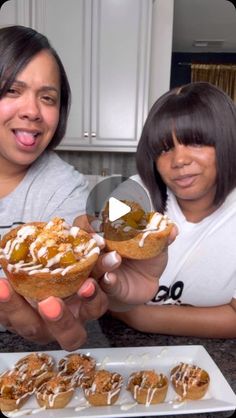 two women holding up small pastries in front of a plate with other desserts on it