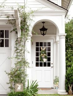 a white house with an arched door and green plants growing on the front porch area