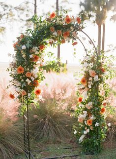 an outdoor wedding arch decorated with orange and white flowers, surrounded by tall grass in the background