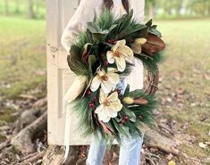 a woman standing in front of a tree holding a wreath with white flowers and greenery