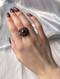 This angular double Bohemian garnet halo ring is just divine! Circa 1970, this piece pulls most of it’s design from the wildly popular bohemian garnet pieces of the 19th century Victorian era; which clustered these bright lustrous garnets together to hi-light their rich color. The double halo of garnets encircle a large central garnet in an almost sunflower-like formation due to the pointed cut of outermost halo, encompassing the flower child era it was created and paying homage to the bohemian garnet jewelry that came before it. A ring perfect for someone who loves statements with subtle hints of color, as the garnets are rather understated until they burst into a bright and brilliant cardinal red in direct light. Materials and Features: Garnet, 9ct Yellow Gold Age/Origin: Vintage, c 1970 Vintage Statement Rings, Vintage Red Garnet Cluster Ring, Vintage Garnet Cluster Ring As Gift, Garnet Ring With 17 Jewels, Vintage Ruby Multi-stone Cluster Ring, Vintage Multi-stone Ruby Cluster Ring, Vintage Garnet Cluster Ring With Gemstone, Vintage Ruby Cluster Ring With Multi-stones, Victorian Ruby Cluster Ring With Gemstones