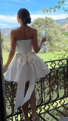 a woman in a white dress standing on a balcony holding a glass of wine and looking out the window