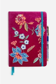 a red notebook with colorful flowers on the front and blue trimmings, sitting next to a pen