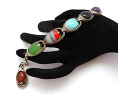 A Vintage faux gemstone set naturalistic adjustable length panel bracelet by Miracle. The bracelet is formed from silver tone metal and mock gemstone set cabochons, including carnelian, onyx,  agate and turquoise.  Fastens with a adjustable length spring ring clasp, complete with ornate drop toggle. Circa  1960's - 70's.     Bracelet is approx 17cm - 21.5cm in length ( unfastened ), height is 1.5cm.     Adjustable length spring ring  clasp.     Signed Miracle to the inside of one panel     Very Vintage Adjustable Bracelets With Stones, Adjustable Multicolor Cabochon Bracelets, Naturalistic Design, 70s Jewelry, Bracelet Vintage, Spring Rings, Onyx, Agate, Silver Tone