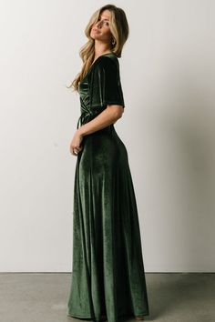 a woman in a green velvet dress standing against a white wall and looking off to the side