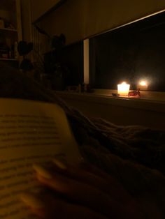 a person reading a book in the dark with two candles lit up behind them on a window sill