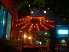 an umbrella is lit up outside at night with lights on it and chairs around the table