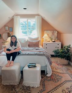 a woman sitting on top of a bed next to a white foot stool in a bedroom