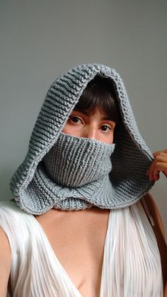 a woman wearing a gray knitted hood and scarf over her face with one hand