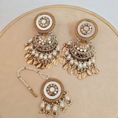 Elevate your bridal look with our Pearl Golden Mirror Jhumka Earring Set, complete with a stunning Mang Tika and Suspended Sheesha Jhumki. This exquisite Indian bridal jewelry is the perfect blend of tradition and style, designed to complement your wedding saree or lehenga. Radiate elegance and sophistication on your special day. Materials: Brass, Pearl, Mirror ❋❋ Shipping Methods ❋❋ Standard Delivery - Take up to 8-14 business days (Worldwide). ❋❋ Please be aware that the colors, shades, and texture shown may exhibit minor variations compared to the actual product due to limitations in digital imaging. We kindly ask for your understanding and consideration of these slight discrepancies. White Wedding Sets With Latkans, Wedding Chandbali Jhumkas For Navratri, Chandbali Jhumkas For Wedding And Navratri, Festive Chandbalis With Gota Work For Reception, Traditional Danglers For Wedding And Navratri, White Tikka With Latkans For Navratri, Bollywood Style Gota Work Danglers For Reception, Traditional Heavy Sharara For Reception, Bridal Earrings With Latkans For Navratri Wedding