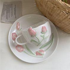 a cup and saucer with pink flowers painted on the side, next to a basket