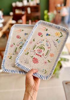 two plates with designs on them being held by a person's hand in front of a dining room table