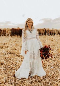 a woman in a white dress standing in a field with cows behind her and holding a bouquet of flowers