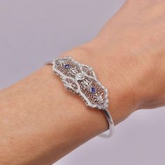 This is part of Chairish’s Fine Jewelry assortment.  A delicate Art Deco bracelet made of 14k white gold with a European cut diamond centre and two French cut sapphires on the side. The centrepiece has a symmetrical design, filled with delicate filagree and a scrolling foliate motif that is detailed and beautifully crafted. The diamond measures 4 mm and has .25 carat weight, it is G/H in colour and VS11 in clarity. The sapphires measure about 2.5 mm and have a well saturated blue tone. The centr Art Deco Jewelry Bracelet, Art Deco White Gold Diamond Cut Bracelet, Art Deco Silver Bracelets With Single Cut Diamonds, Art Deco Silver Diamond Bracelet, Art Deco Silver Diamond Cut Bracelet, Art Deco Hallmarked White Gold Bracelets, Saturated Blue, Symmetrical Design, French Cut