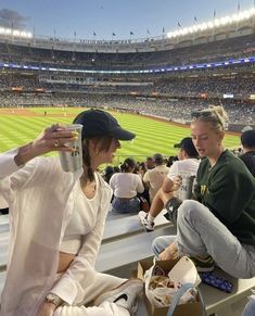 two women sitting on the bleachers at a baseball game eating and drinking beverages