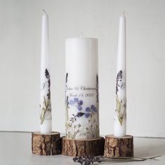 three white candles with blue flowers on them sitting next to each other in front of a white wall