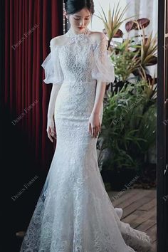 a woman standing in front of a window wearing a wedding dress with an open shoulder