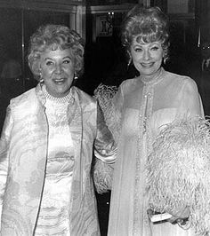 two women standing next to each other in front of a building wearing evening dresses and fur stolers