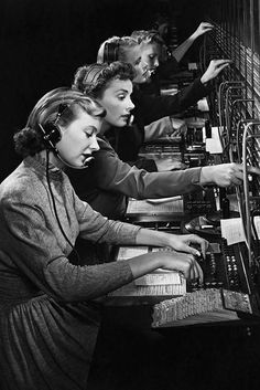 three women in headphones are working on an old fashioned computer screen with many rows of keyboards