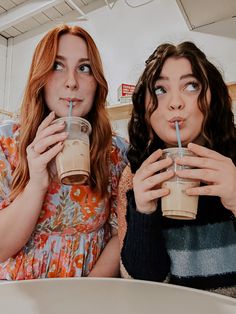 two women sitting at a table with drinks in their hands and one holding a straw