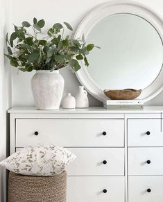 a white dresser topped with a round mirror next to a vase filled with greenery