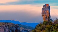 a tall rock tower sitting on top of a lush green hillside next to trees and mountains