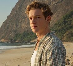 a young man standing on top of a sandy beach next to the ocean with mountains in the background