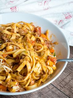 a white bowl filled with pasta and meat on top of a wooden table next to a fork