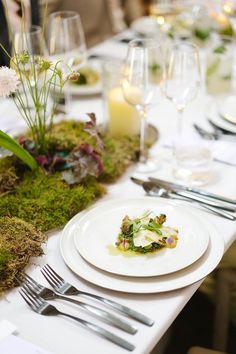 the table is set with white plates and silverware, moss covered placemats