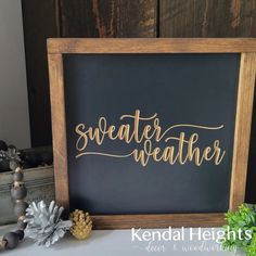 a wooden frame with the words sweater weather written in gold on it next to some pine cones