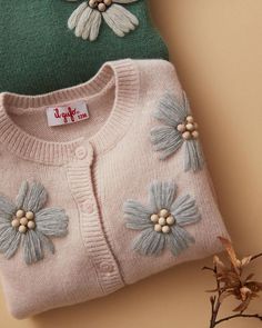 two knitted sweaters with flowers on them sitting next to each other, one is pink and the other is green