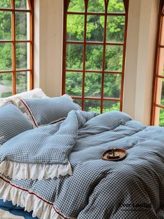 an unmade bed with blue and white checkered bedspread in front of two windows