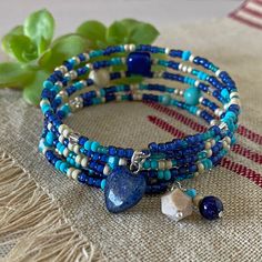 "Notice how the glass seed beads sparkle and shimmer in the video clip! And, I love these beautiful shades of blue all mixed with an accent of creamy beige in a type of bracelet that I wear quite often - memory wire!  Both the sparkle and matte finish of the seed beads add texture to this 5-wrap bracelet that is enhanced by the three focal beads - a tumbled Lapis nugget, a soft Riverstone round, and a lovely \"Goldline\" Turquoise (dyed Magnesite). The seed beads include a rich royal blue, soft matte turquoise, silver lined teal, and exquisite Duracoat matte galvanized silver.  The Swarovski crystal bi-cone beads include \"Golden Shadow\", \"Dark Sapphire\", and turquoise.  A few smaller Goldline Turquoise rounds are scattered throughout the bracelet as well.  The charms at one end of the Blue Spiritual Wrap Bracelet With Gemstone Beads, Spiritual Blue Gemstone Beads Wrap Bracelet, Spiritual Blue Wrap Bracelet With Gemstone Beads, Turquoise Heart-shaped Beaded Bracelet, Blue Gemstone Beads Wrap Bracelet, Blue Gemstone Beads Wrap Bracelet With Round Beads, Turquoise Heart-shaped Beaded Bracelets, Blue Beaded Bracelets With Heart Beads, Blue Beaded Heart Bracelet