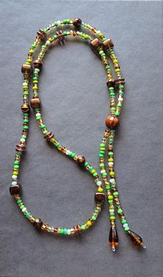 Tassel beaded necklace made with green/gold/brown/pearlescent seed beads, various shaped brown glass beads, and a ceramic accent bead leading to two beaded tassels with tear drop beads on the ends. The necklace measures 16" long and slides over the head (no clasp). Makes a great statement necklace for layering or by itself! Adjustable Brown Beaded Necklaces With Oval Beads, Adjustable Brown Beaded Necklace With Dangling Beads, Bohemian Brown Beaded Necklaces With Oval Beads, Brown Beaded Necklaces With Oval Beads, Brown Beaded Chain For Festival, Brown Beaded Necklace With Colorful Oval Beads, Brown Double Strand Beaded Necklace For Gift, Adjustable Green Beaded Necklace With Wooden Beads, Artisan Green Beaded Necklace With Dangling Beads