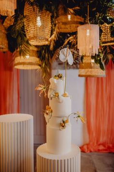 a three tiered white wedding cake with flowers on top and hanging lights in the background