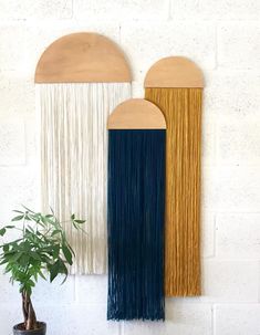 three different colored wall hangings on a white brick wall, one with fringe and the other with wood