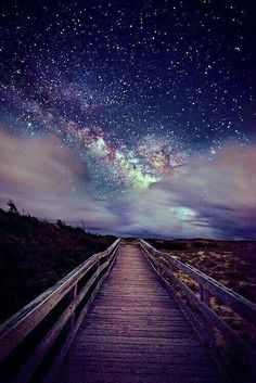 the night sky is filled with stars above a wooden walkway leading to an open field