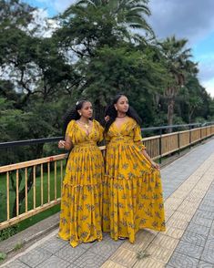 two women in yellow dresses standing next to each other on a bridge with trees in the background