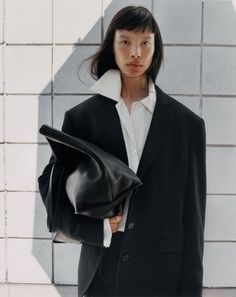 a woman in a black suit and white shirt is holding a black bag while standing next to a brick wall