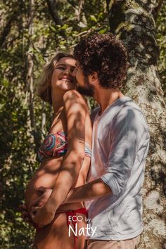 a man and woman standing next to each other in front of a tree with their arms around each other