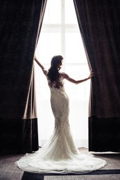 a woman standing in front of a window wearing a wedding dress and holding her arms out