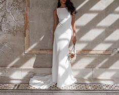 a woman standing in front of a wall wearing a white dress and holding a bouquet