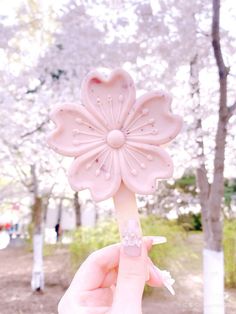 a hand holding a pink flower shaped object in front of some trees and flowers on the ground