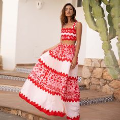 a woman in a red and white dress standing on steps near a cactus tree with her hands behind her back