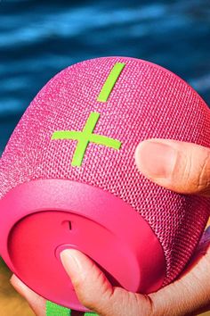 a person holding a pink speaker with green crosses on it's side, near the water