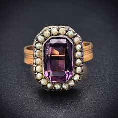 "Amethyst stones have been thought for generations to provide protection and were worn by the Ancients as a talisman for luck. The beautiful violet purple hue has also had an allure not to be resisted. Beloved by Victorians as well, it was the first color a widow was allowed to wear after coming out of mourning.  This ring was converted from an antique brooch. The beautiful, faceted rectangular amethyst is haloed by 21 natural seed pearls, which the Victorian jewelers implemented to represent \"tears of sorrow\". The faceted emerald cut amethyst stone is paired with a replica textured band, giving its new incarnation as a ring is a lovely way to showcase this classic combination of amethyst and pearl.   Details (approximate) Size: 7 Weight:  5 grams Materials: Gold, Amethyst, Seed Pearl Me Heirloom Amethyst Ring With Center Stone, Purple Amethyst Gemstone With Center Stone, Hallmarked 14k Gold Purple Amethyst Ring, 14k Gold Hallmarked Purple Amethyst Ring, Heirloom Purple Amethyst Ring With Center Stone, Heirloom Purple Rings With Accent Stones, Heirloom Purple Ring With Center Stone, Heirloom Purple Amethyst Ring For Formal Occasions, Amethyst Ring With 17 Jewels For Anniversary