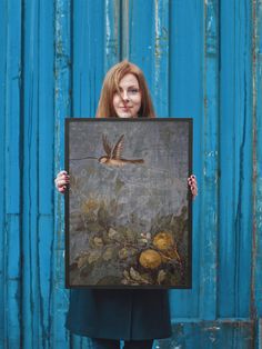 a woman holding up a painting in front of a blue wall with a bird on it