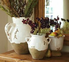 two white vases with flowers and fruit in them on a wooden table next to a mirror