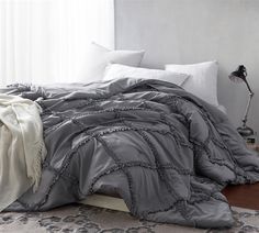 an unmade bed with grey and white comforter on top of it next to a window