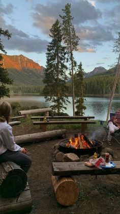 three people sitting around a campfire in the woods with mountains in the back ground